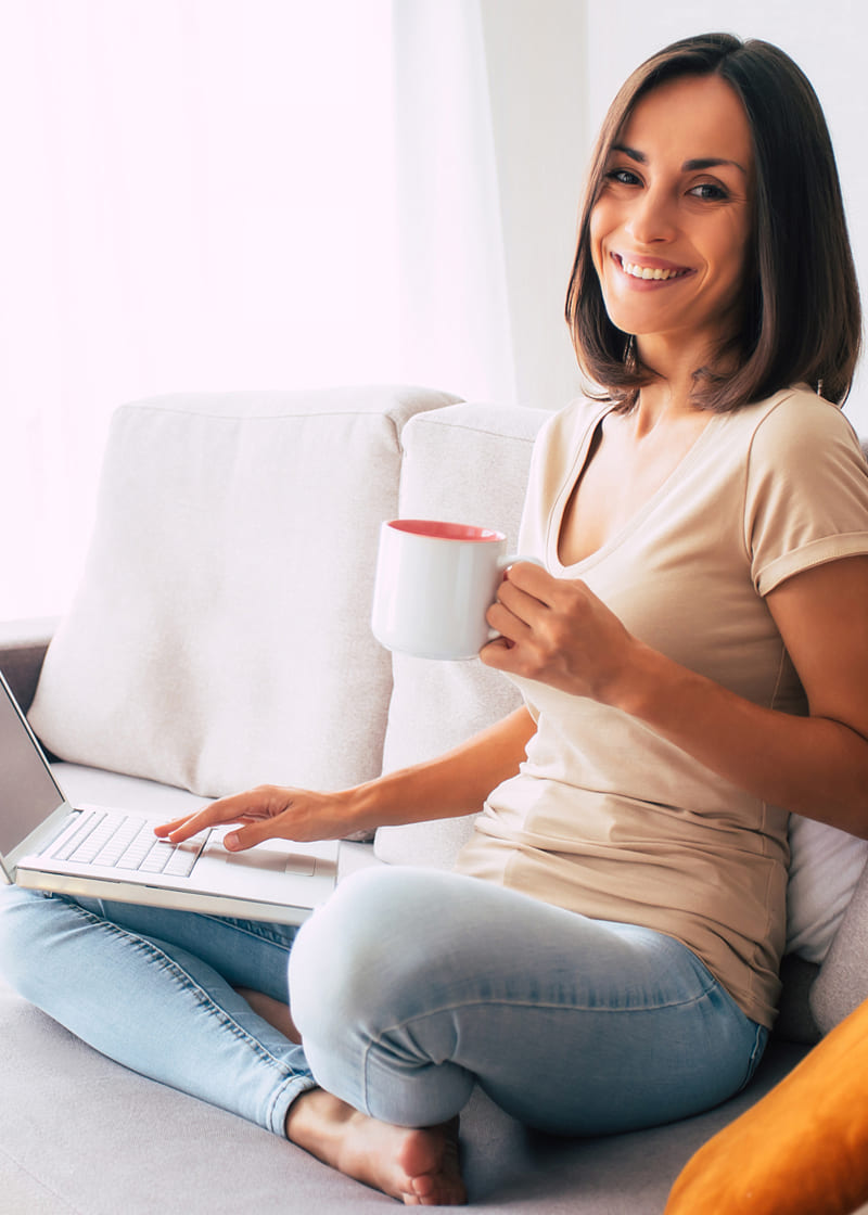 Tânără pe o canapea cu cafea și laptop<br />
